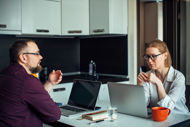 Domowe biuro podczas samoizolacji. Mąż i żona w okularach siedzą przy stole w kuchni, pracują razem zdalnie na laptopach, dyskutują i uśmiechają się. Niezależny rodzinny.