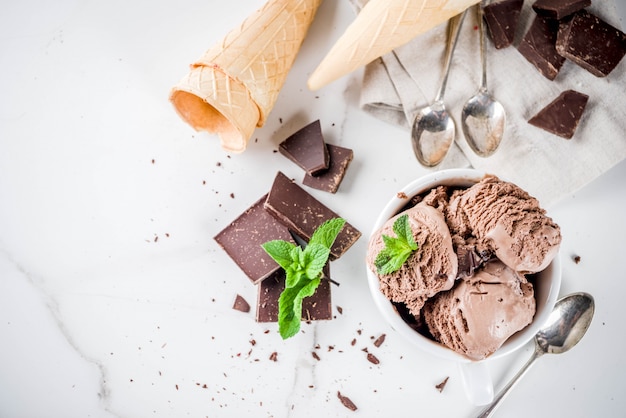 Domowe lody czekoladowe podawane ze świeżą miętą, ciemną czekoladą i rożkami lodów waflowych, powyżej biały marmurowy stół