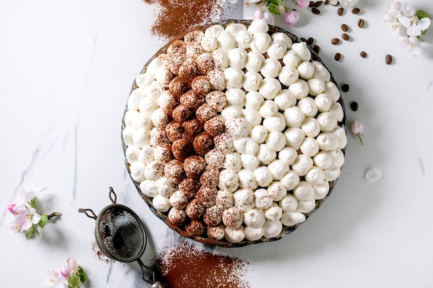 Domowy bezglutenowy tradycyjny włoski deser tiramisu posypany kakao w proszku ozdobiony kwitnącą jabłoń i ziarnami kawy na białej marmurowej powierzchni. Widok z góry, układ płaski. Skopiuj miejsce