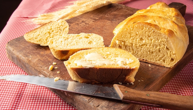 Domowy chleb, krojony domowy chleb z masłem na drewnie i obrusem w czerwono-białą kratkę, nóż i pęczek pszenicy.