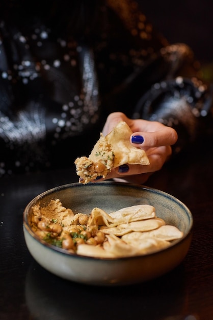 Domowy hummus ozdobiony ciecierzycą, pietruszką, czosnkiem i oliwą z oliwek Kobieca dłoń maczająca trójkąty z chleba pita w misce hummusu