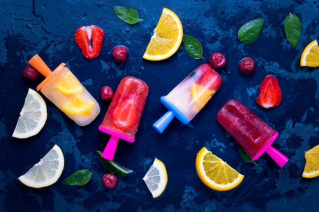 Domowy popsicle jasnych owoców o smaku truskawkowym, wiśniowym, cytrynowym, pomarańczowym, cytrynowym i miętowym oraz świeżych owoców na lody na ciemnoniebieskim