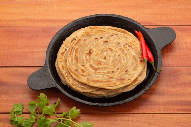 Zdjęcie domowy porotta lub parathawarstwowy płaski chleb z mąki pszennej ułożony w żeliwnej patelni lub thawie z czerwonym chilli i liściem kolendry na drewnianym stole na białym tle