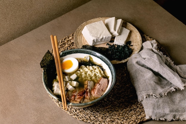 Domowy ramen w stylu azjatyckim z makaronem grillowana pierś z kaczki tofu wodorosty nori i jajko na twardo w ceramicznej misce z pałeczkami i łyżką na brązowym stole kuchennym