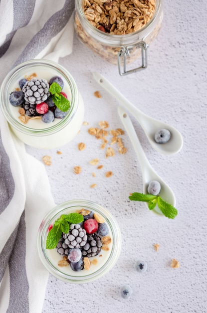 Domowy świeży jogurt w szklanym słoju z mrożonymi jagodami i muesli na wierzchu. Zdrowe śniadanie. Orientacja pionowa.