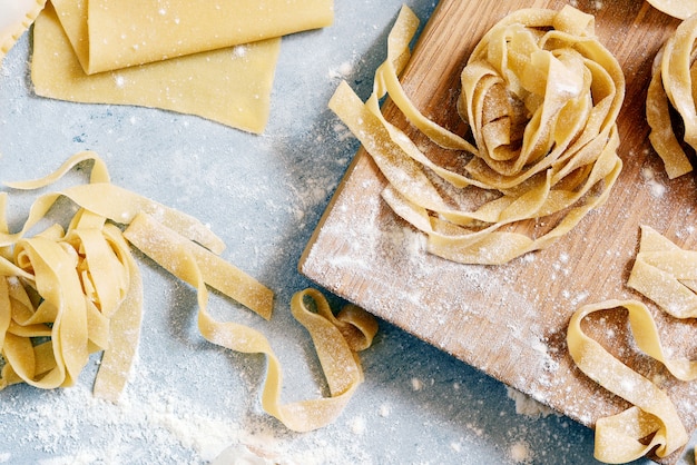 Domowy włoski makaron, ravioli, fettuccine, tagliatelle na drewnianej desce i na niebieskim tle. Proces gotowania, surowy makaron