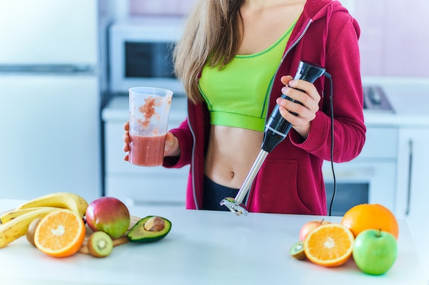 Dopasowana Kobieta W Stroju Sportowym Przygotowuje W Domu świeży, Organiczny Koktajl Owocowy Za Pomocą Ręcznego Blendera W Kuchni.