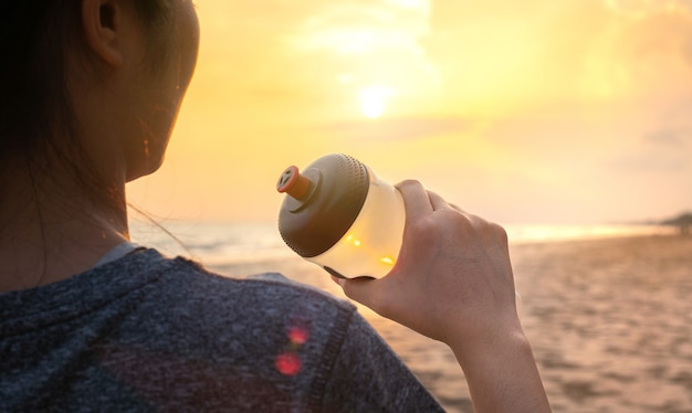 Dopasuj młoda kobieta pije świeżą wodę na relaks i odpoczynek po biegu, ćwiczenia sportowe na plaży, morze w piękny zachód słońca