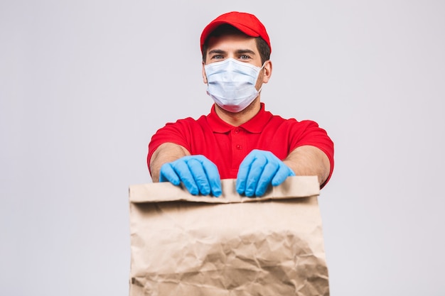Doręczeniowego mężczyzna pracownik w czerwonej nakrętki koszulki munduru twarzy maski pustych rękawiczkach trzyma pustego karton odizolowywającego na biel ścianie. Usługa kwarantanna pandemiczny wirus koronawirusa koncepcja 2019-ncov.