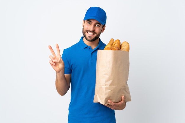 Doręczeniowy mężczyzna trzyma torbę pełno chleby odizolowywający na biel ścianie uśmiecha się zwycięstwo znaka i pokazuje