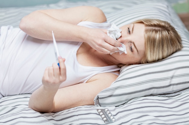 Zdjęcie dorosła biała kobieta leżąca w piżamie w łóżku, sprawdzająca temperaturę na termometrze i dmuchająca nos w chusteczkę. wiele wzorów.