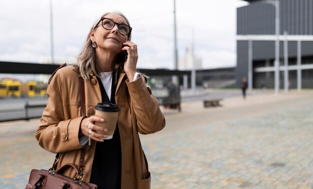 Dorosła biznesowa kobieta rozmawia przez telefon na zewnątrz z filiżanką kawy