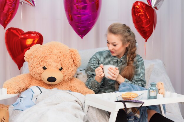 Dorosła dziewczyna pije herbatę z misiem zabawka w łóżku na Walentynki