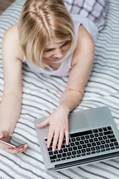 Dorosła kobieta w białej piżamie, leżąca na kanapie na brzuchu, surfuje po Internecie za pomocą laptopa, trzymając w ręku smartfon. Widok z góry. Pionowy.
