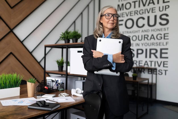 Dorosła kobieta w biznesowym stroju stoi z laptopem w dłoniach na tle