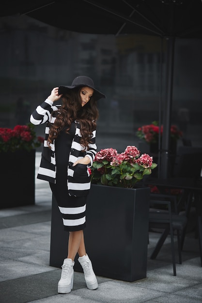 Dorosła kobieta w modnym czarnym kapeluszu i modnych białych butach pozuje na europejskiej ulicy miasta
