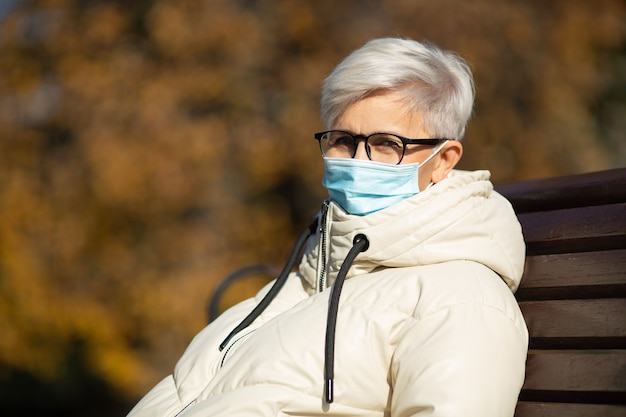 dorosła kobieta w wieku siedząca na ławce w parku jesienią ubrana w maskę medyczną