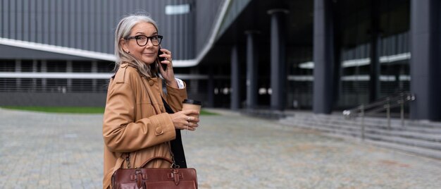 Dorosła, Odnosząca Sukcesy Kobieta Biznesu Wchodzi Do Budynku Biurowego Rozmawiając Przez Telefon Komórkowy Z Filiżanką