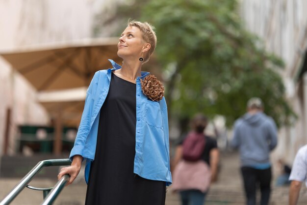 Zdjęcie dorosła stylowa uśmiechnięta kobieta o krótkich blond włosach stojąca na europejskiej ulicy w jasnoniebieskim kolorze