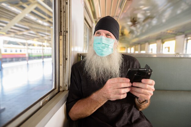 Dorośleć brodatego turysty z maską dla ochrony przed epidemią wirusa korony myślenie podczas korzystania z telefonu wewnątrz pociągu