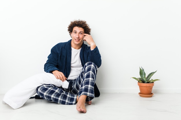 Dorośleć mężczyzna jest ubranym piżamę siedzi na podłogowej domu pokazuje rozczarowanie gest z palcem wskazującym.