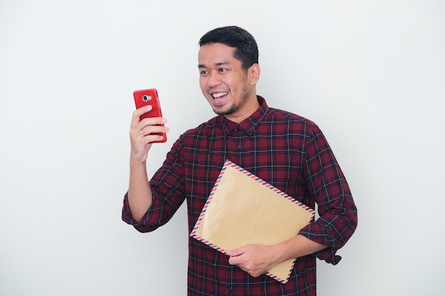 Dorosły Azjata trzymający kopertę pocztową z wyrazem szczęśliwej twarzy, gdy czyta wiadomość na swoim telefonie komórkowym