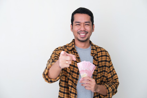 Dorosły Azjata trzymający papierowe pieniądze, wskazując palcem do przodu i uśmiechnięty