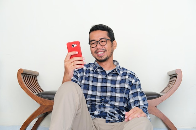 Dorosły azjatycki mężczyzna siedzący zrelaksowany, patrzący na swój telefon komórkowy ze szczęśliwym wyrazem twarzy