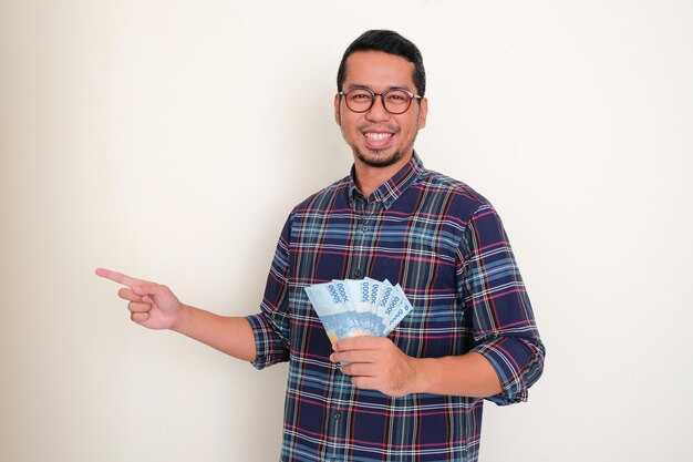 Dorosły azjatycki mężczyzna uśmiecha się i wskazuje prawą stronę, trzymając pieniądze