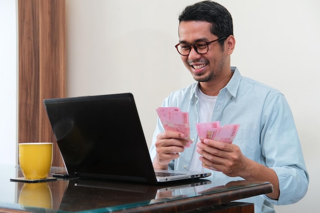Dorosły azjatycki mężczyzna uśmiecha się szczęśliwy, licząc swoje pieniądze przed laptopem