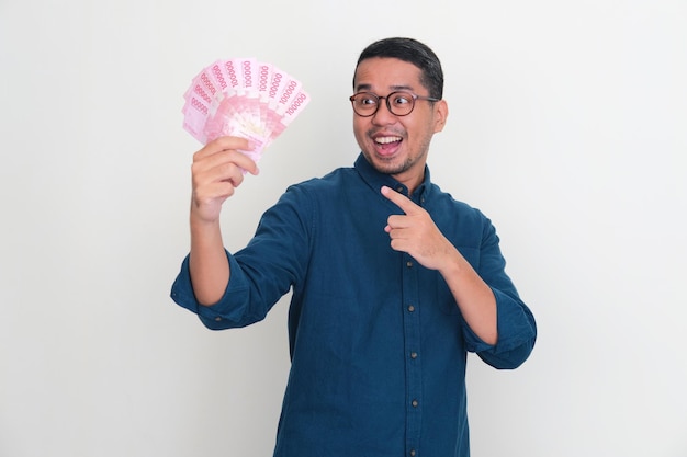 Dorosły azjatycki mężczyzna z podekscytowanym wyrazem twarzy, wskazujący na pieniądze, które trzyma