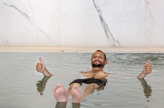 Dorosły kaukaski mężczyzna z brodą wąsami unoszący się w basenie z gęstą słoną wodą i pokazujący podwójny kciuk w górę ok tak Brodaty mężczyzna unoszący się w basenie spa z wodą z Morza Martwego