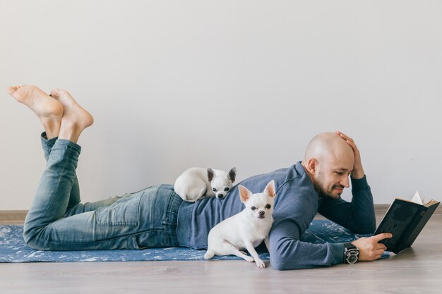 Dorosły łysy mężczyzna w modnej odzieży leżącej na macie jogi. Młody chłopak, czytanie książki. Dwa białego szczeniaka chihuahua w domu