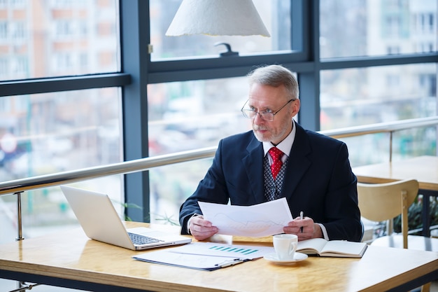 Dorosły mężczyzna mentor, reżyser, biznesmen w okularach i garniturze studiujący dokumenty siedząc przy stole. Koncepcja dnia roboczego