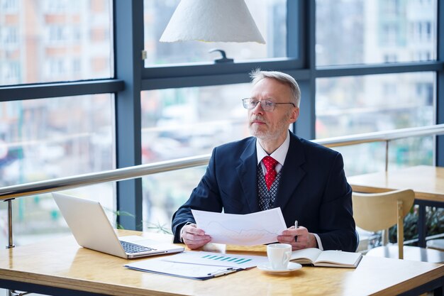 Dorosły mężczyzna mentor, reżyser, biznesmen w okularach i garniturze studiujący dokumenty siedząc przy stole. Koncepcja dnia roboczego