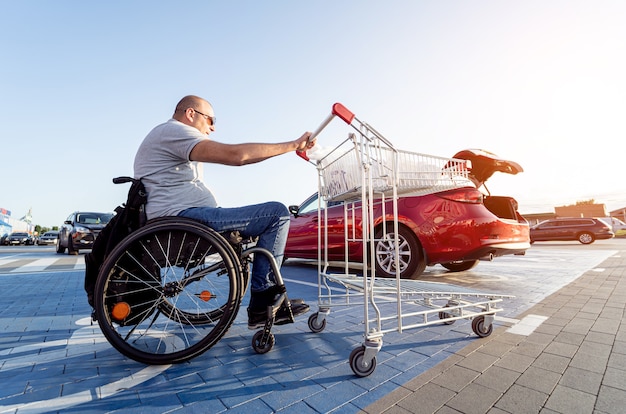 Dorosły niepełnosprawny mężczyzna na wózku inwalidzkim pcha wózek w kierunku samochodu na parkingu supermarketu