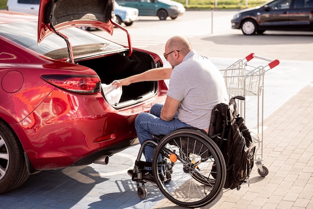 Dorosły niepełnosprawny mężczyzna na wózku inwalidzkim wkłada zakupy do bagażnika samochodu na parkingu supermarketu