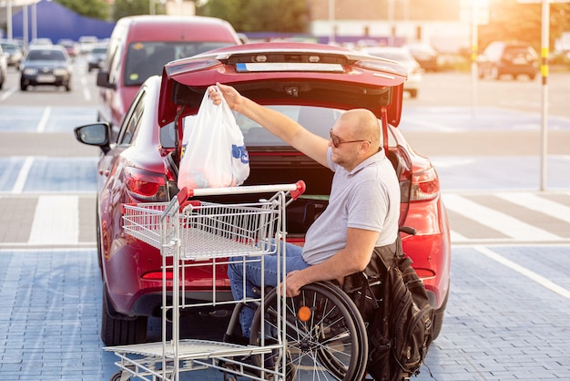 Zdjęcie dorosły niepełnosprawny mężczyzna na wózku inwalidzkim wkłada zakupy do bagażnika samochodu na parkingu supermarketu