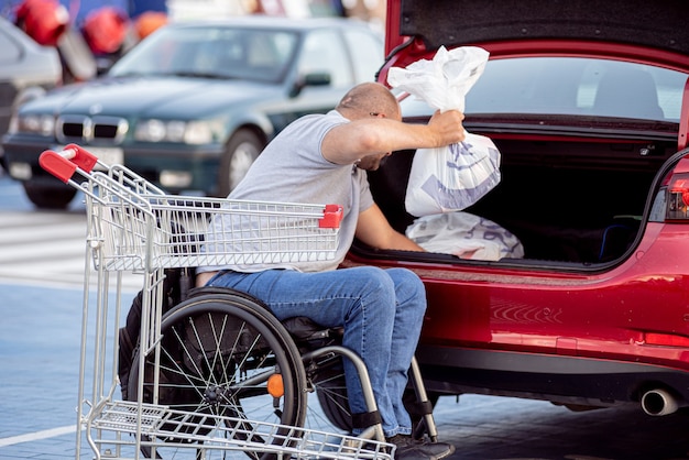 Dorosły niepełnosprawny mężczyzna na wózku inwalidzkim wkłada zakupy do bagażnika samochodu na parkingu supermarketu