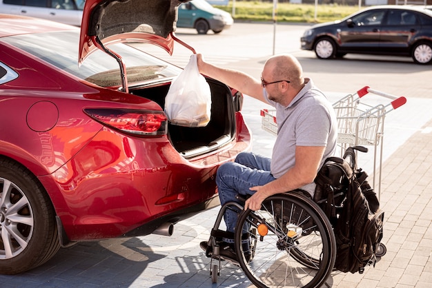 Dorosły niepełnosprawny mężczyzna na wózku inwalidzkim wkłada zakupy do bagażnika samochodu na parkingu supermarketu