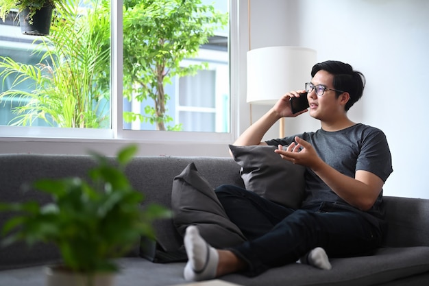Dorywczo azjatycki człowiek siedzi na wygodnej kanapie i rozmawia przez telefon komórkowy.