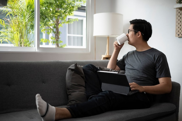 Dorywczo azjatycki człowiek siedzi na wygodnej kanapie w domu i pije kawę rano.