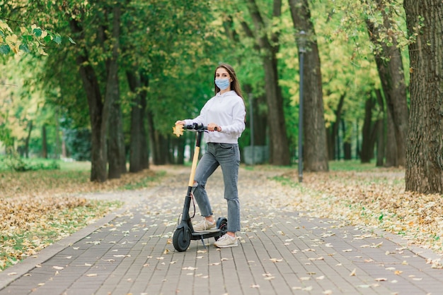 Dorywczo kaukaski kobieta nosi maskę ochronną na miejski skuter elektryczny w parku miejskim podczas pandemii chrząszczy. Koncepcja mobilności miejskiej.