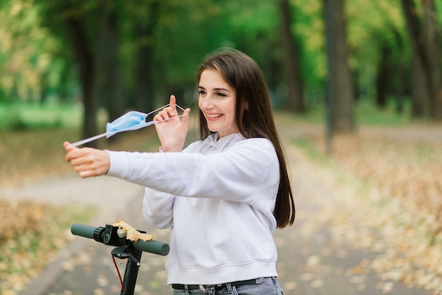 Dorywczo kaukaski kobieta nosi maskę ochronną na miejski skuter elektryczny w parku miejskim podczas pandemii chrząszczy. Koncepcja mobilności miejskiej.