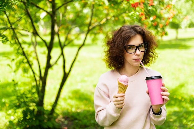 Dość Młoda Kaukaska Kobieta Idzie Na Spacer Do Parku, Pije Kawę I Je Lody