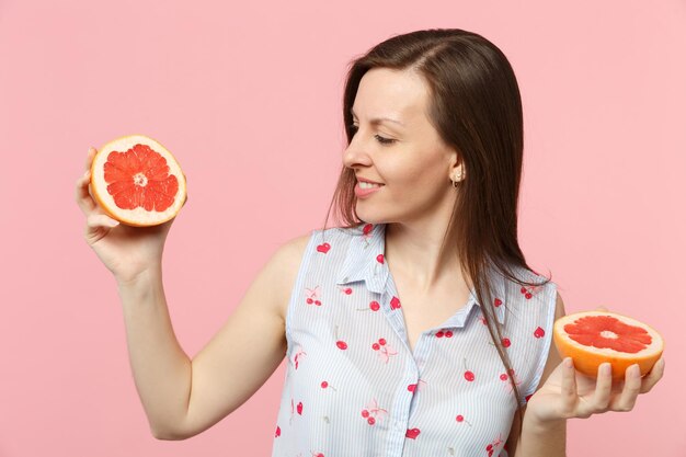 Dość młoda kobieta w letnie ubrania, trzymając dwie połówki świeżych dojrzałych grejpfrutów na białym tle na różowym tle ściany pastelowe w studio. Ludzie żywy styl życia zrelaksować koncepcję wakacji. Makieta miejsca na kopię.