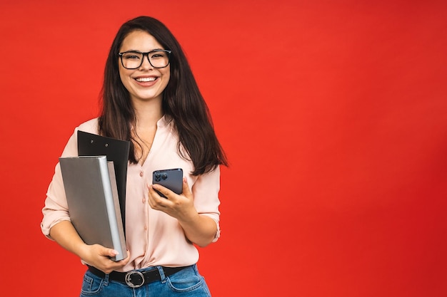 Dość młoda kobieta w swobodnej ręce trzyma laptopa w biurze na białym tle na czerwonym tle Korzystanie z telefonu komórkowego
