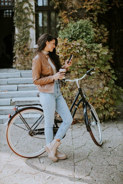 Dość młoda kobieta z telefonem komórkowym pije kawę, aby jechać rowerem w jesienny dzień