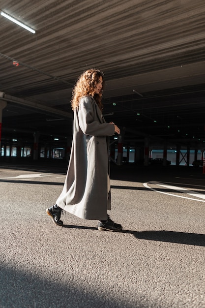 Dość młoda stylowa kobieta z kręconymi włosami w modzie długi płaszcz spacery po ulicy na parkingu. Miejski styl kobiecy