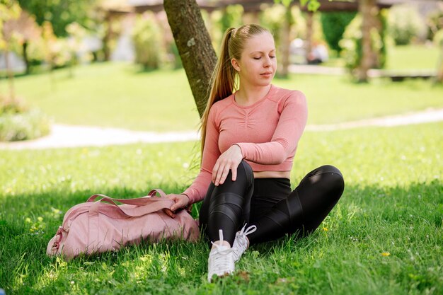 Dość szczupła blond kobieta w sportowej odzieży i sportowej torbie, siedząca i odpoczywająca na trawniku w parku